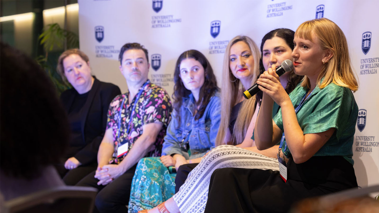 A panel is sitting in front of a white media wall. The woman on the far right is speaking into a handheld microphone.
