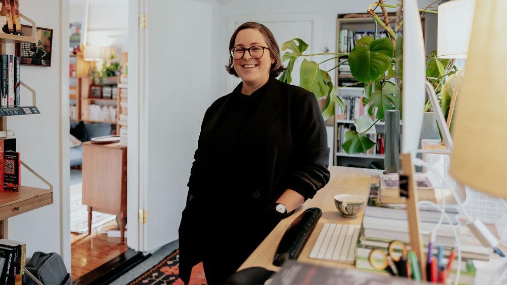 A woman with short brown hair and glasses is standing in her home office, smiling