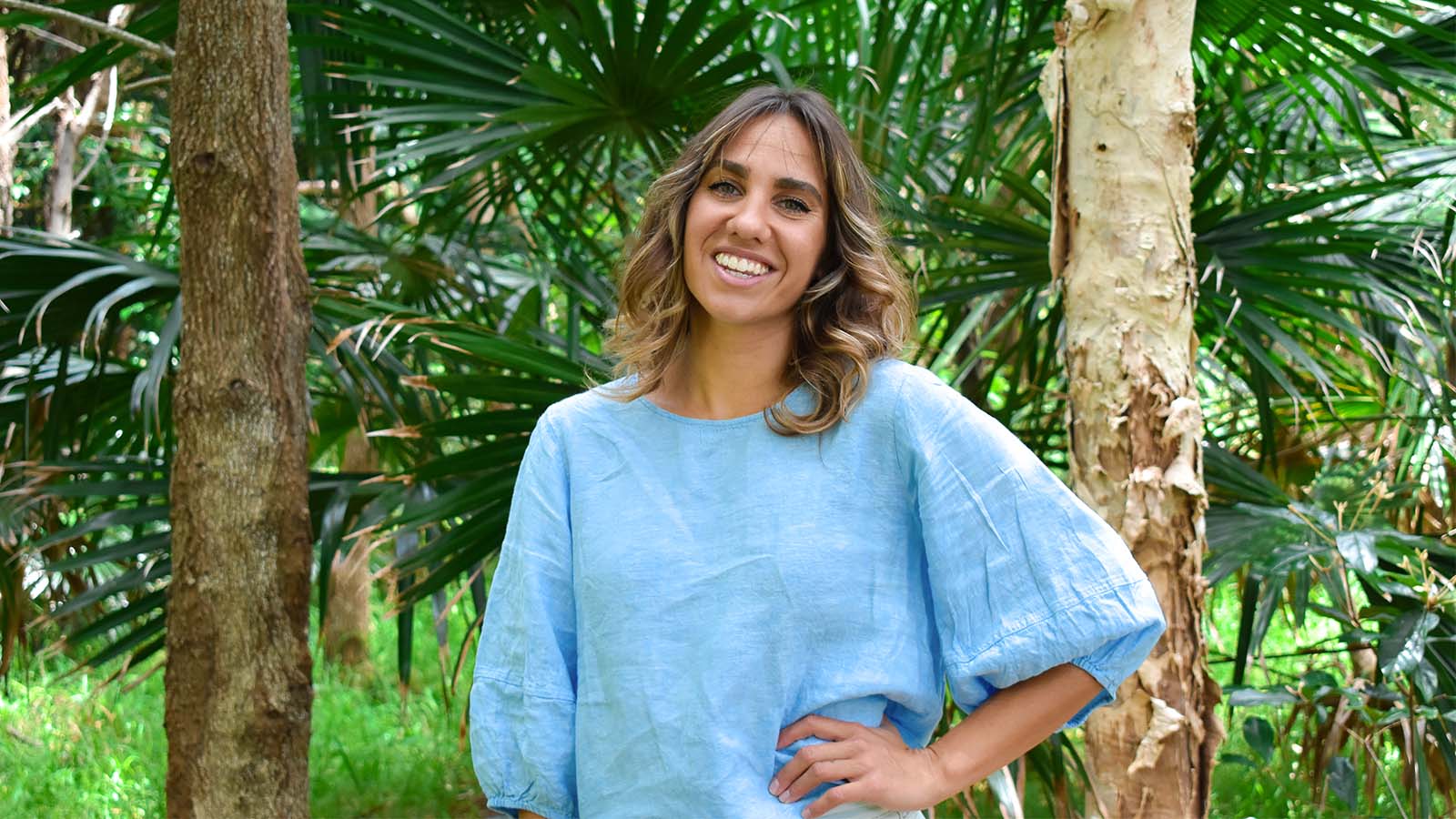 A woman with light brown hair is wearing a bright blue top. She is standing in front of a green bush with one hand on her hip, smiling