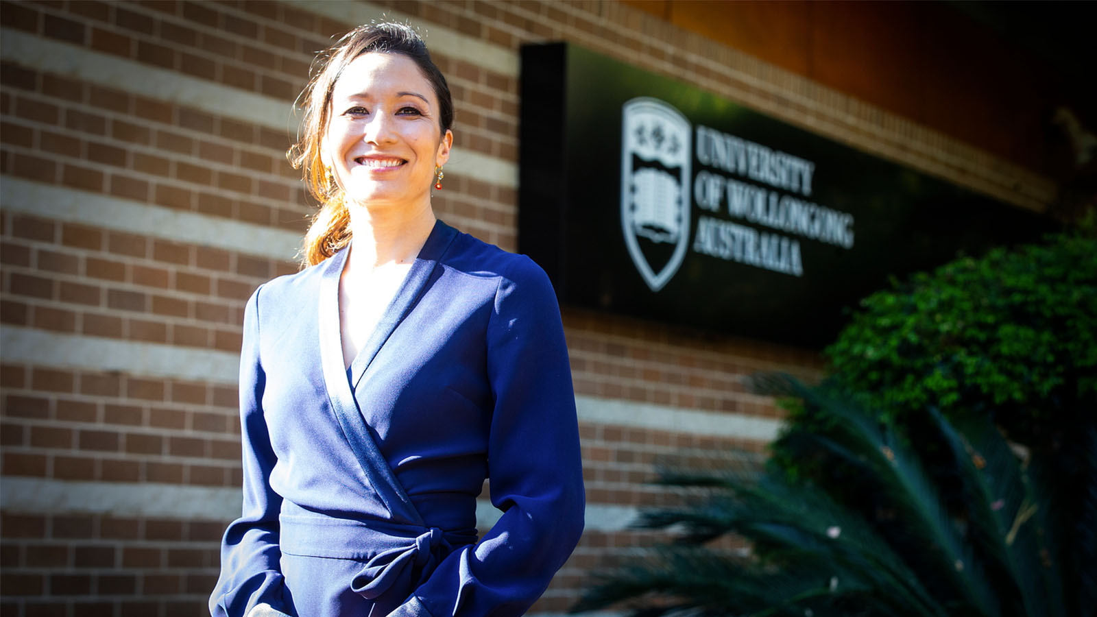 A woman in a blue dress is standing in front of a University of Wollongong Australia sign, smiling