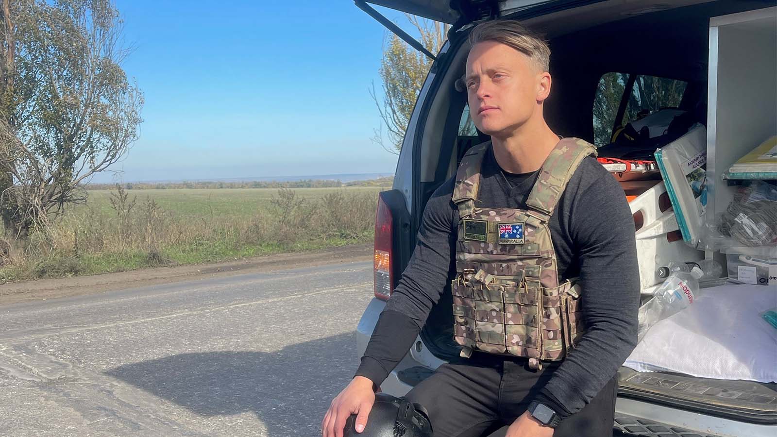 A man with an army vest is sitting in the back of a ute looking into the distance