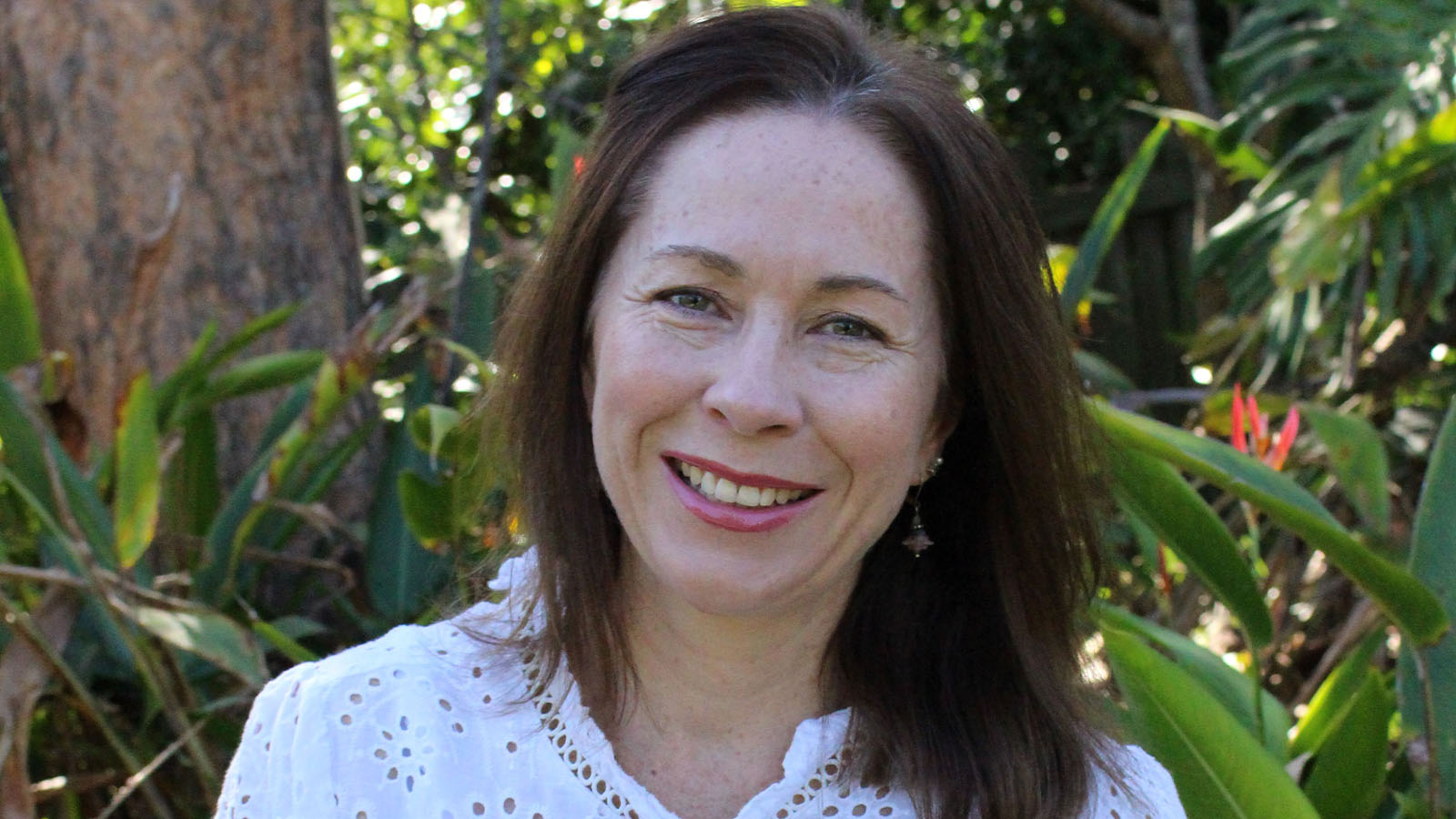 A woman with dark hair is standing in front of a green tree, smiling