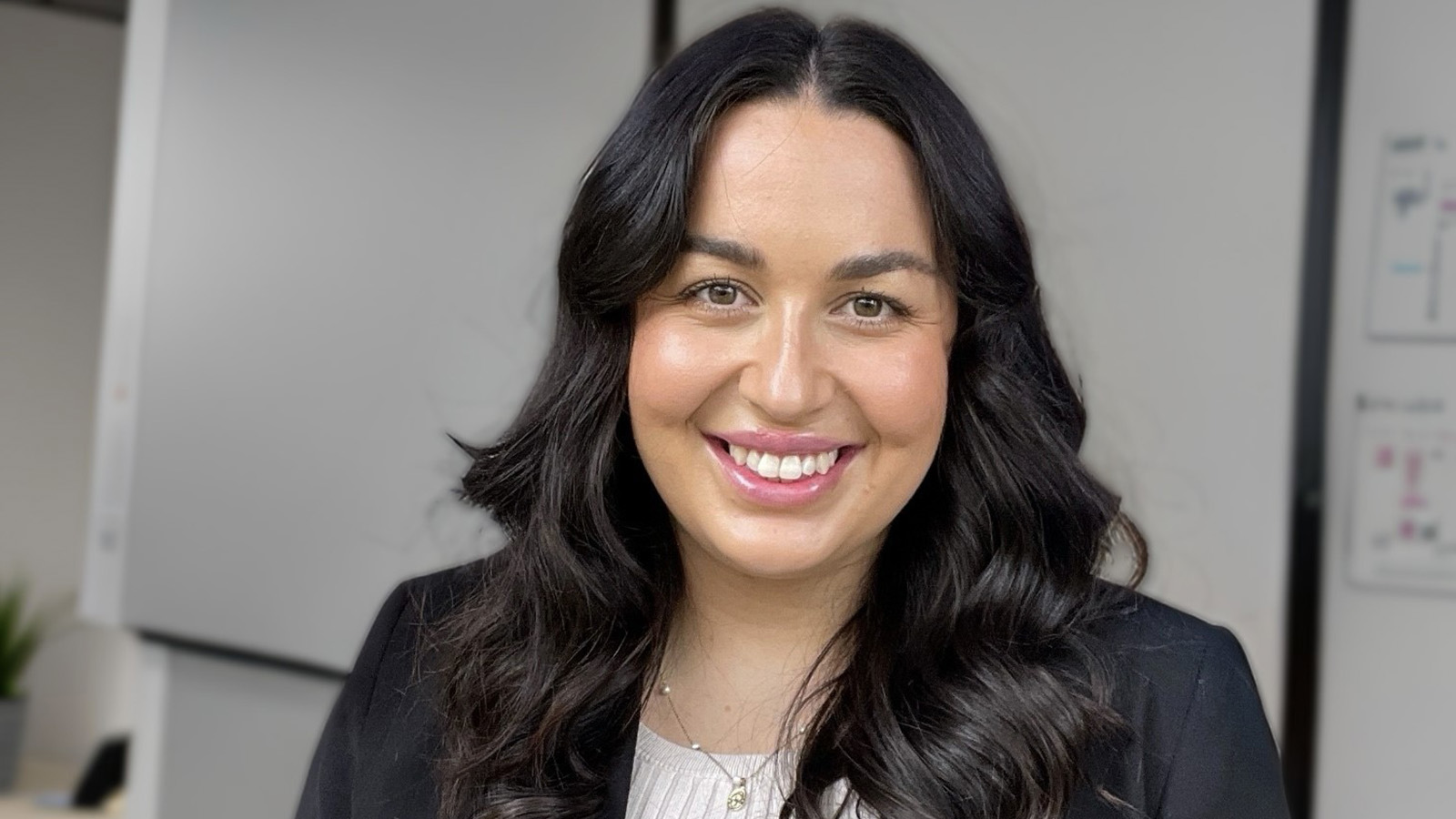A woman with dark hair is wearing a white top and black blazer. She is standing in an office, smiling.