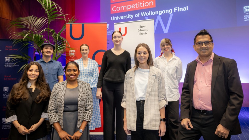 Eight PhD candidates stand on a stage in front of a banner that says 'three minute thesis'