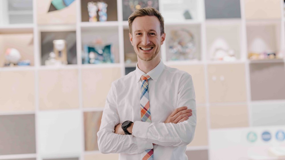 A smiling man in a shirt and tie stands with arms folded.