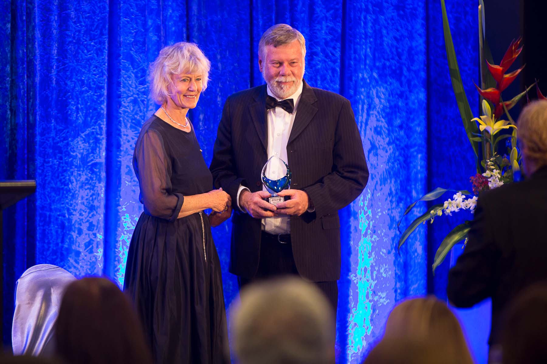 On a stage against a blue backdrop, a woman presents a man with a trophy.