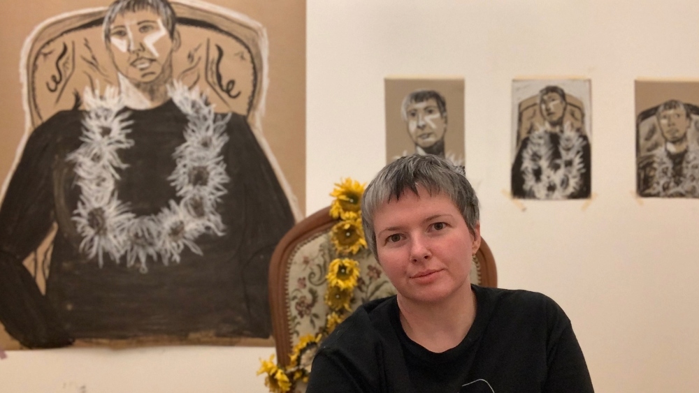 A woman sits facing the camera, a necklace made of flowers hanging from the back of her chair. Stuck to the wall behind are four charcoal self-portraits complete with sunflowers.