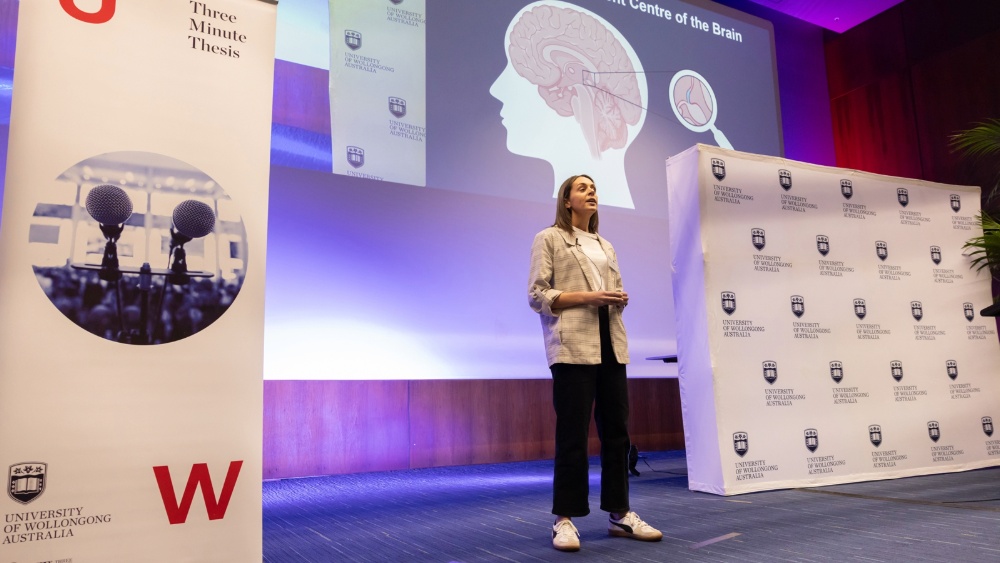 A woman presenting on a stage. An image of a brain is projected behind her with the title 