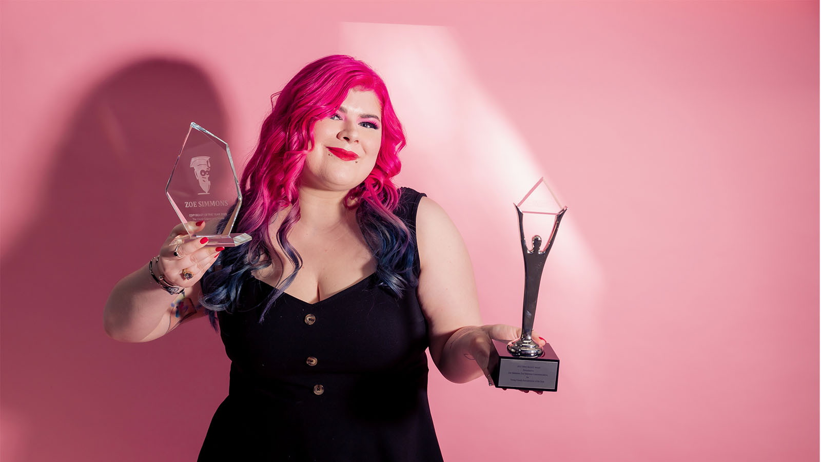 A woman with pink hair and a black dress is standing against a pink backdrop. She is holding two silver awards.