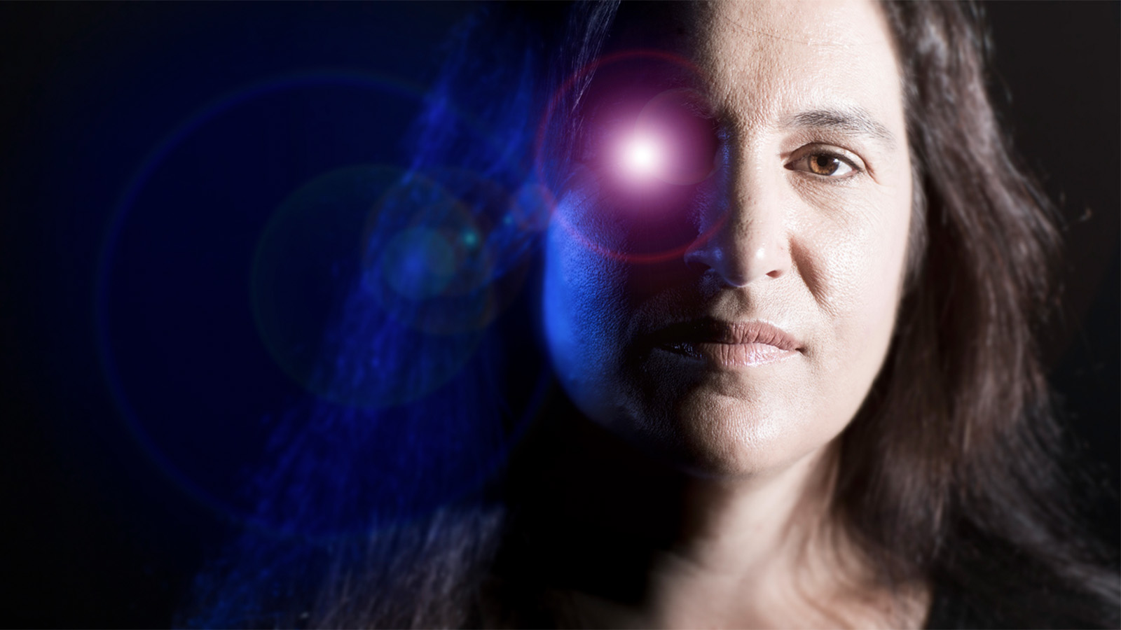 A woman is standing in a dark room with a blue light projected on her. Her right eye is covered by a red light.