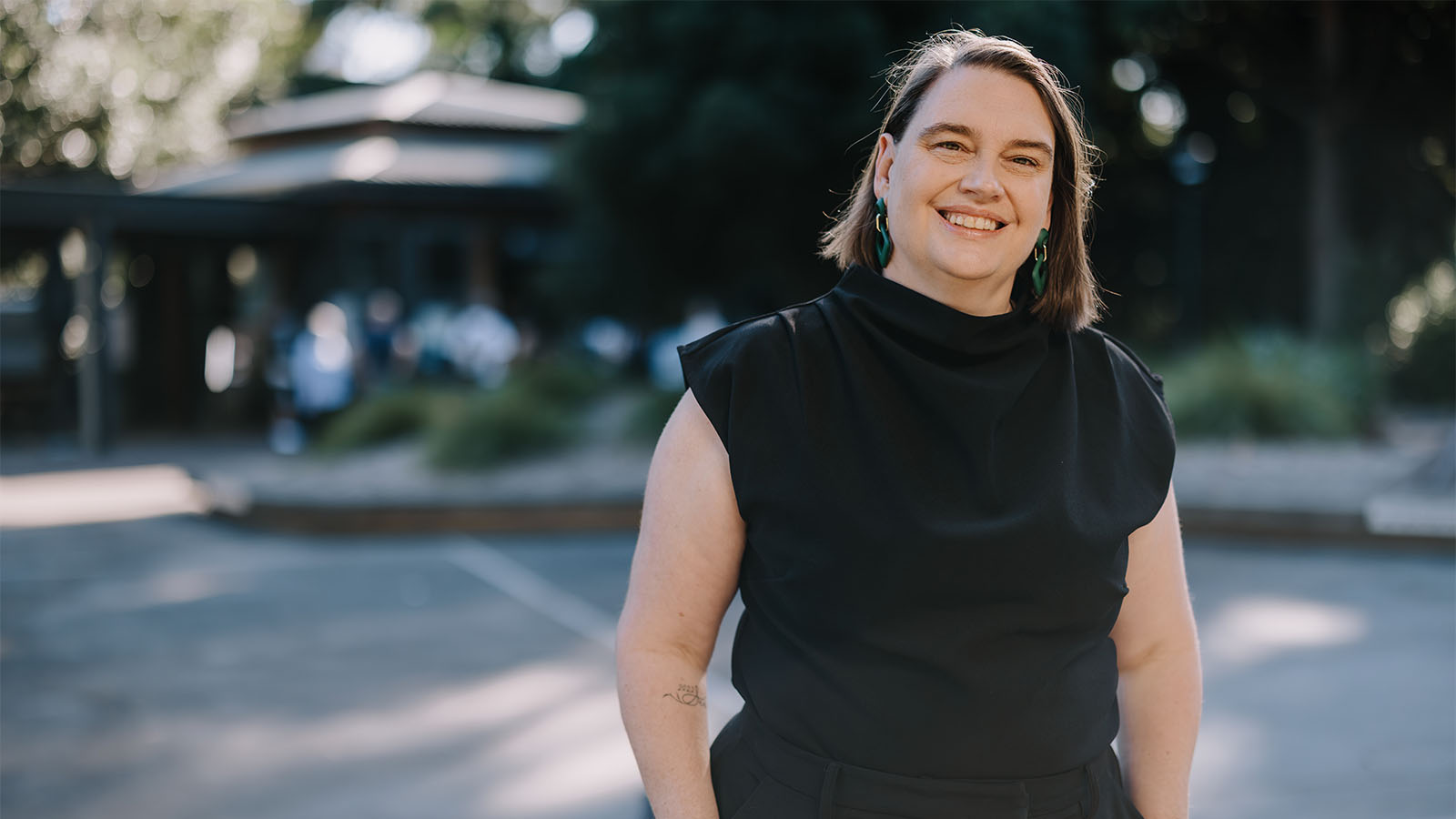 A woman in all black is standing on a basketball court with her hands in her pant pockets, smiling.