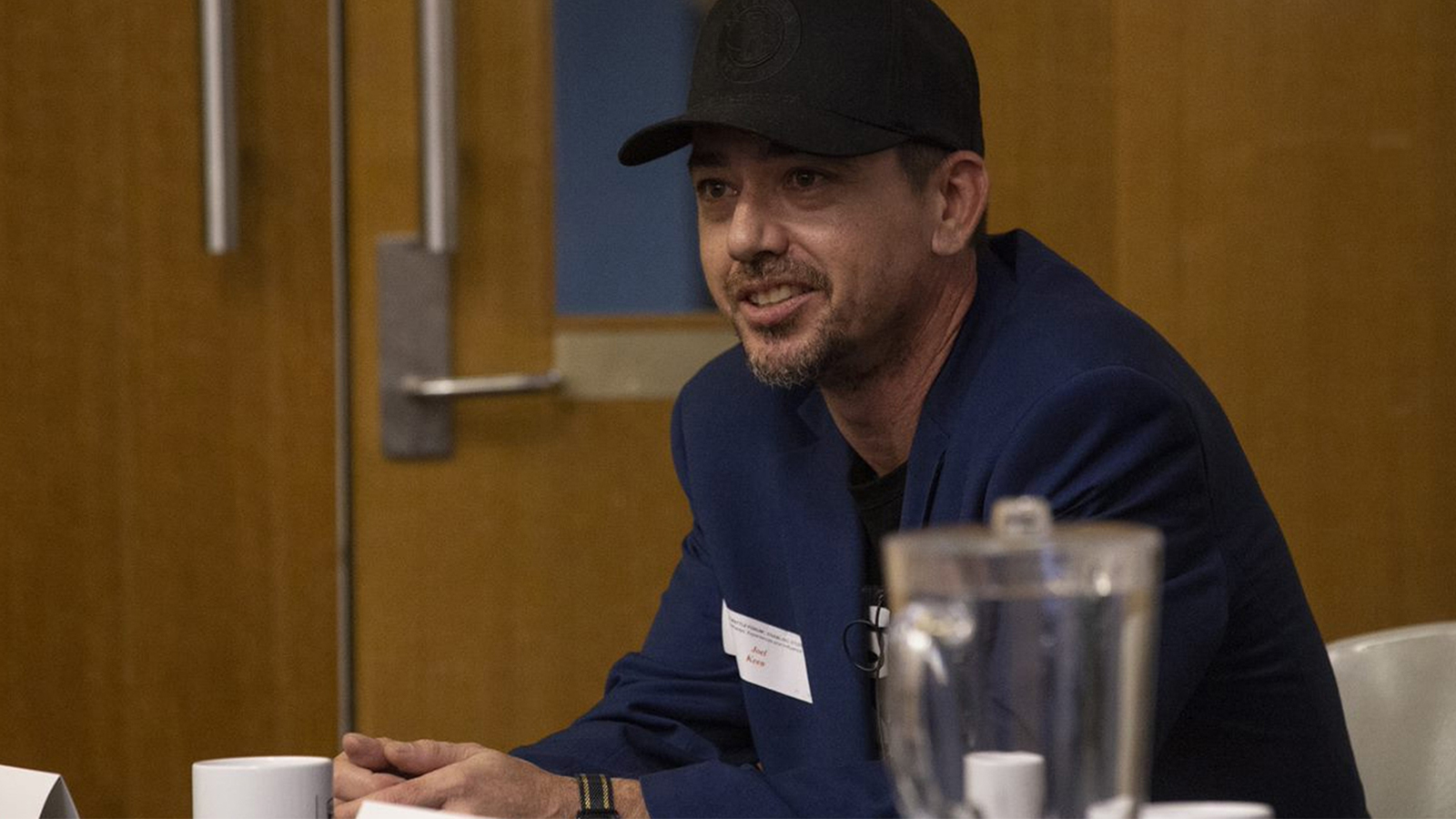 An man in a blue shirt and black cap sitting at a desk in a coference, smiling
