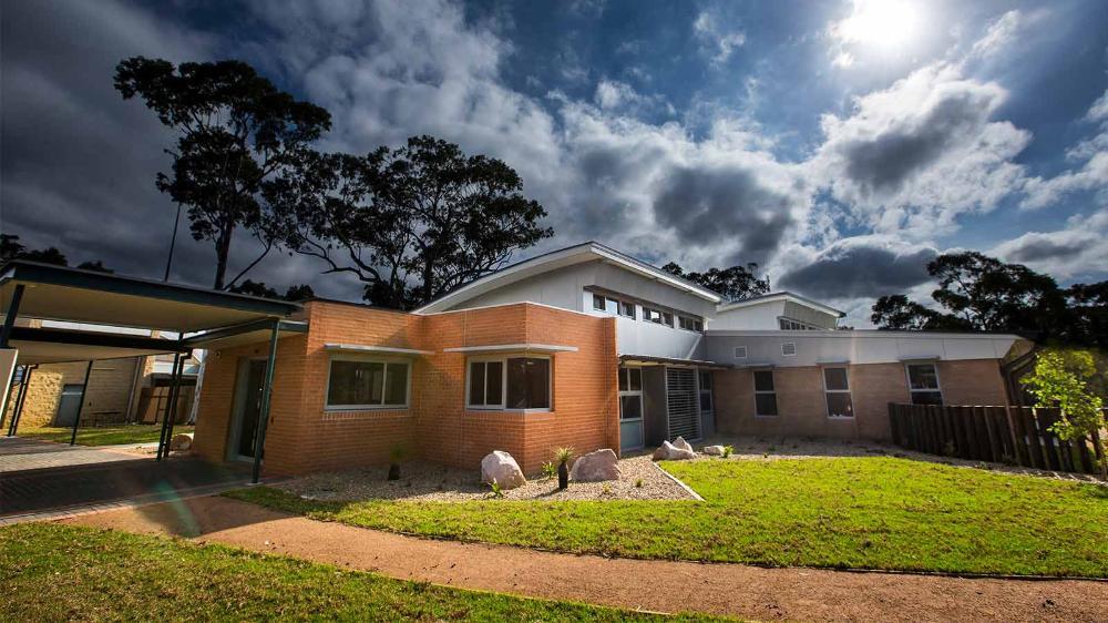 A building at UOW Shoalhaven. It is blonde and orange brick with angled rooves and a small garden.