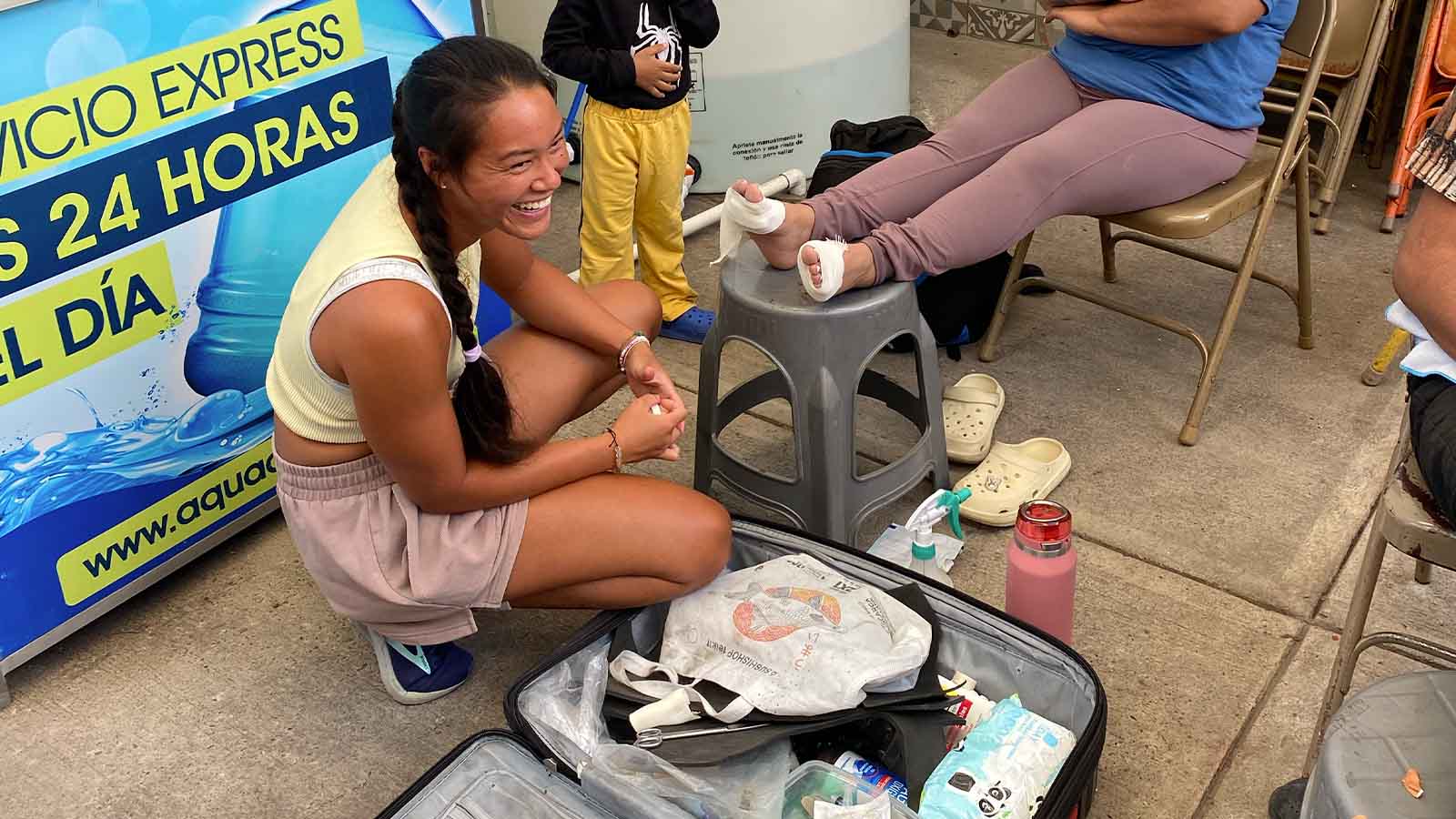 A woman is crouched on the floor next to a medical bag, bandading a patient's feet