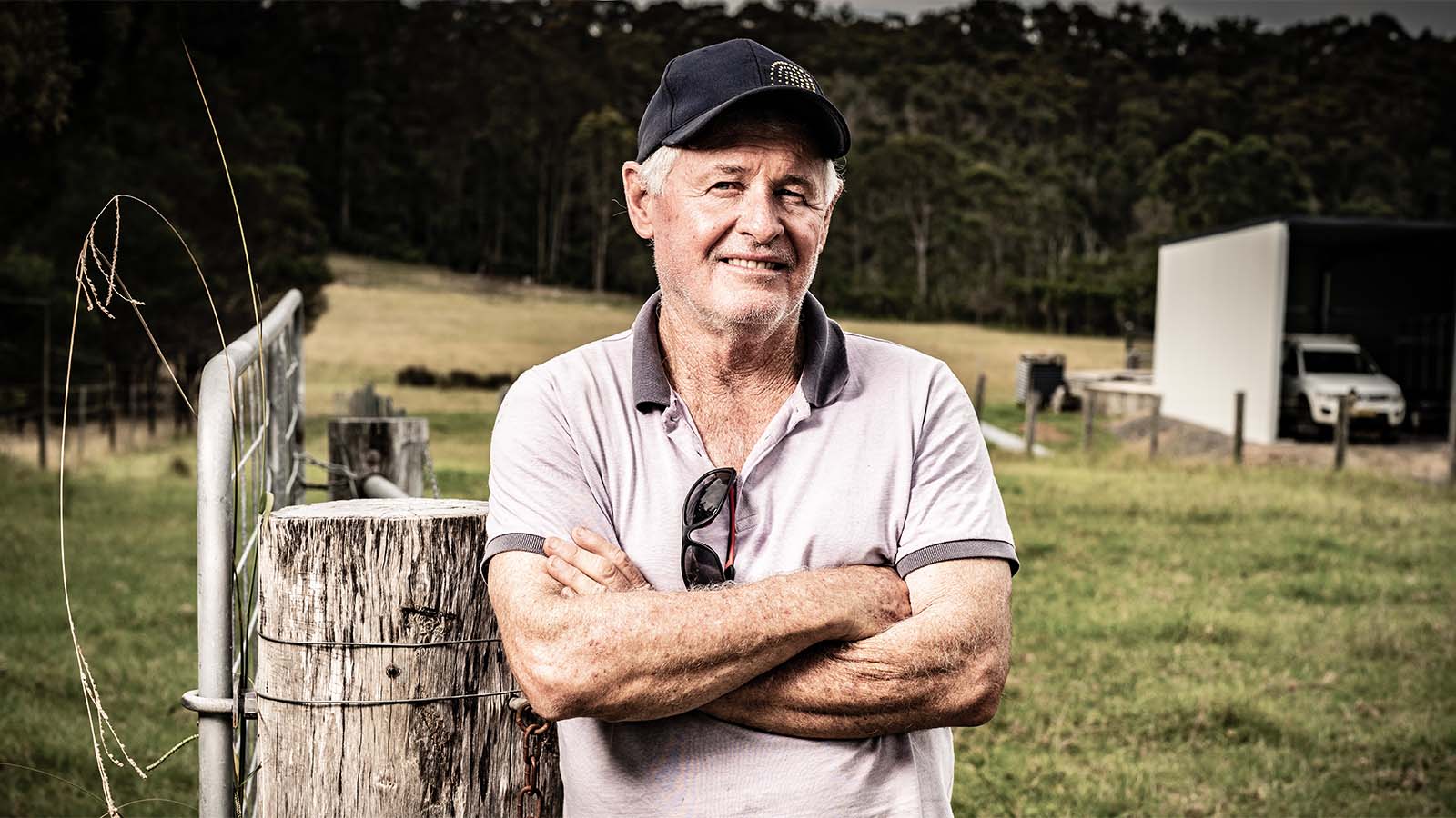 A man in a grey polo shirt and black cap is standing against a farm gate with his arms folded
