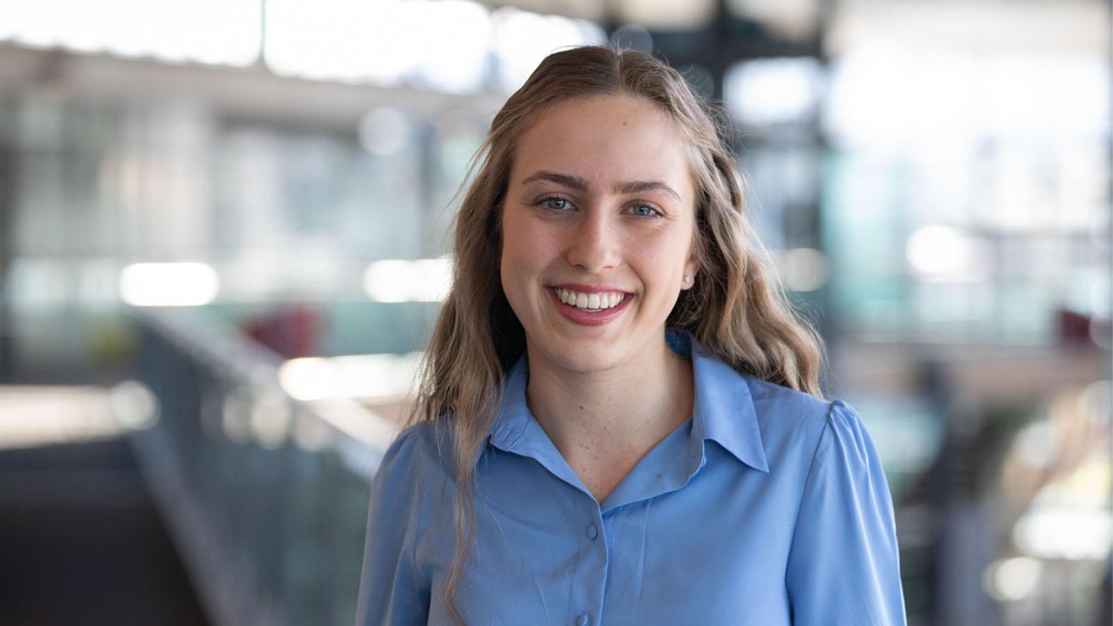 A young woman in a light blue collared business shirt is smiling