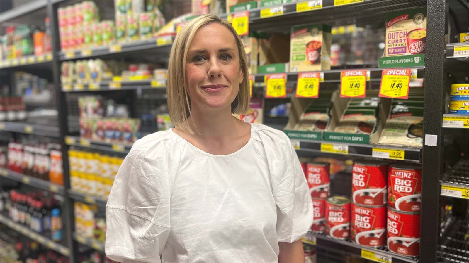 A woman is standing in the aisle of a supermarket in front of tinned vegetables and soups