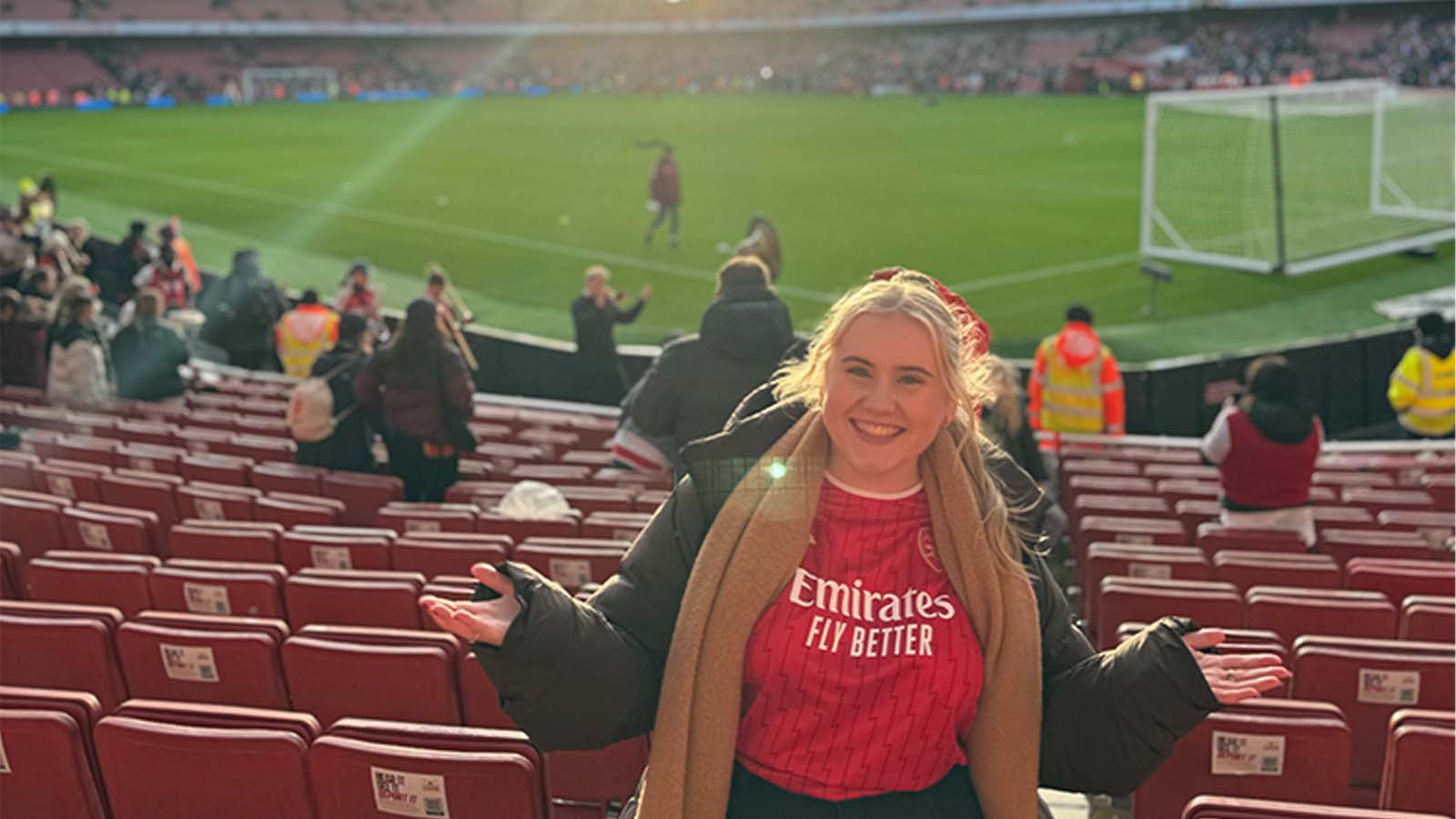 A woman is standing in a football stadium, wearing a red Emirates football jersey, black jacket and brown scarf