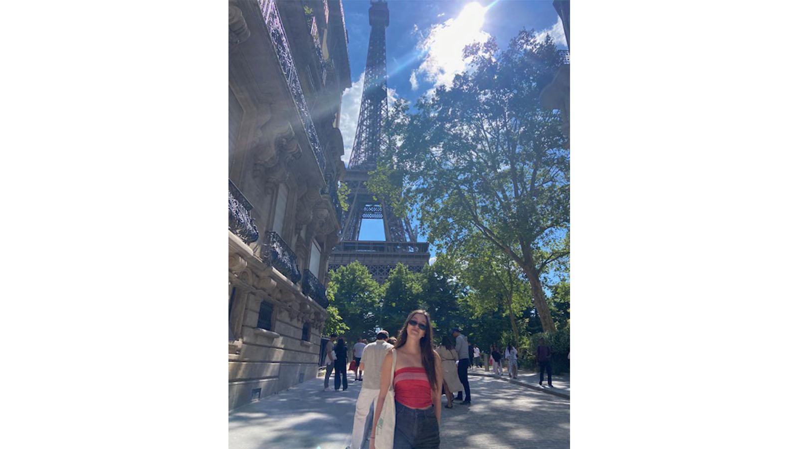 A woman in a red top and blue jeans is standing on a footpath in front of the Eiffel Tower