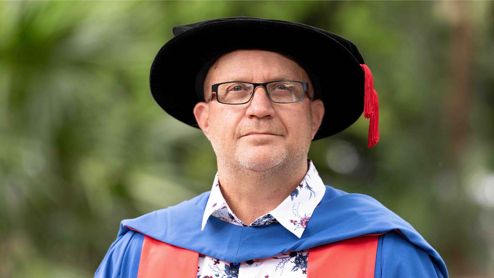 A man with glasses wearing a blue graduation gown and black, floppy graduation hat