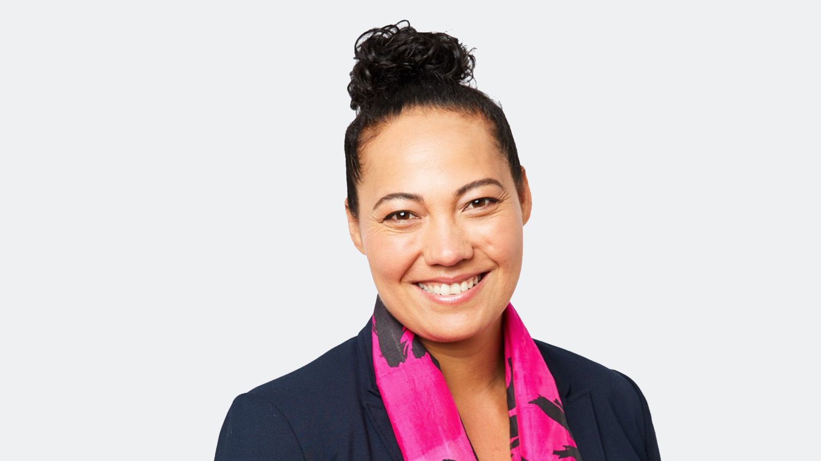 A woman with dark brown hair is standing against a white backdrop, smiling