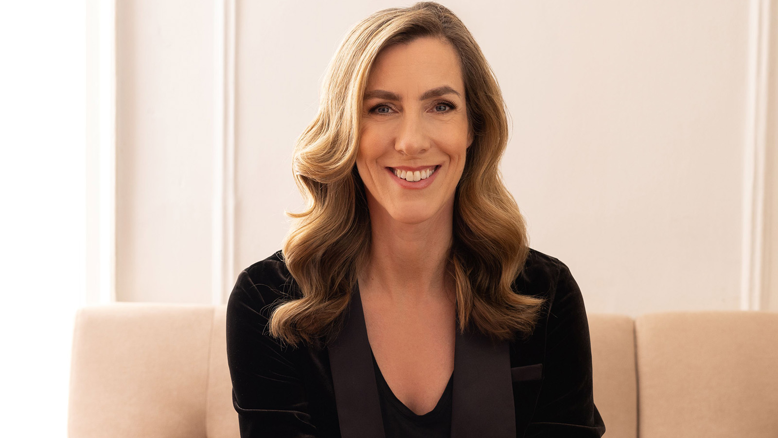 A woman with long light brown hair is sitting on a couch in an office, smiling