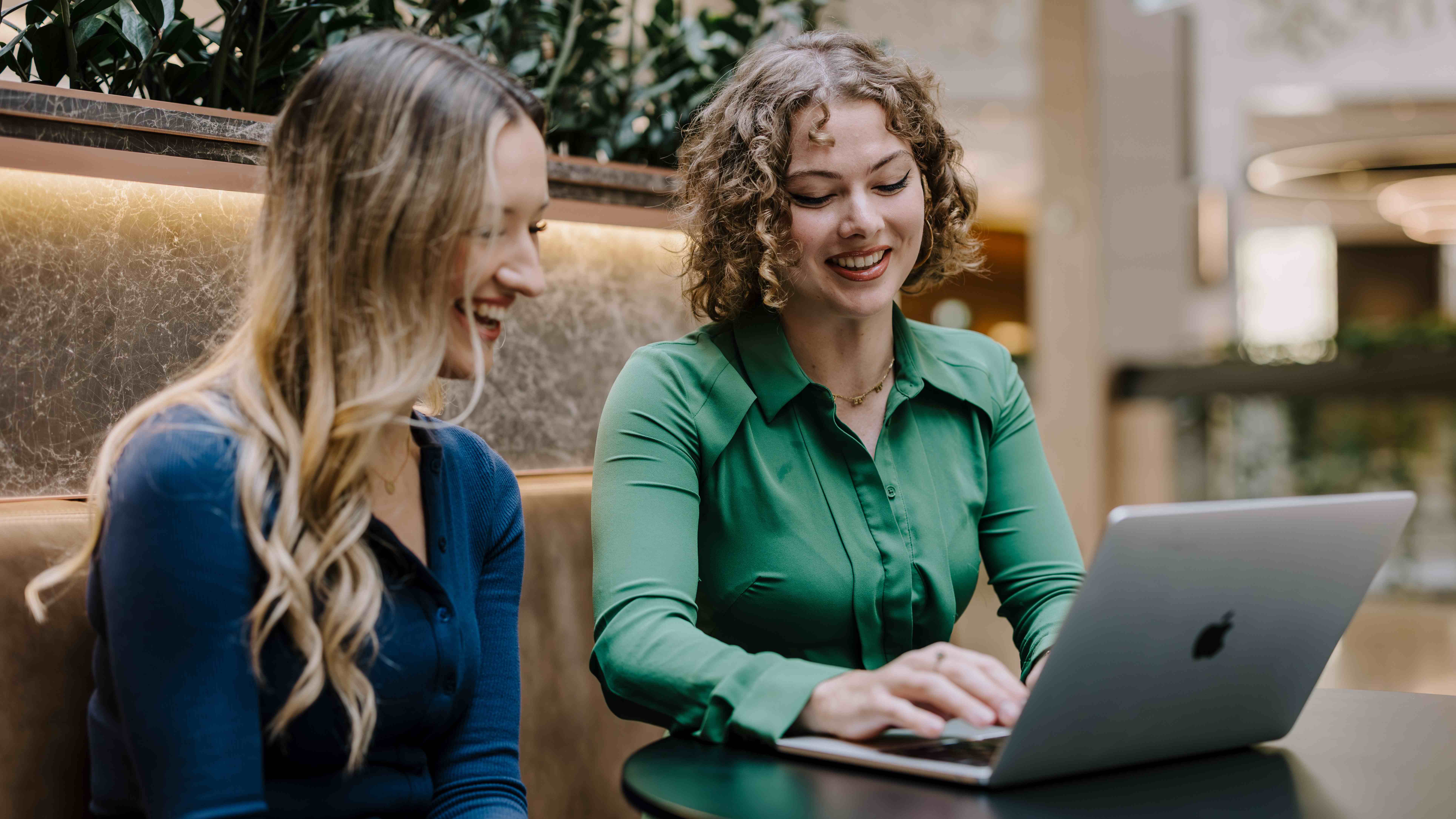 Two female presenting people sitting at a table indoors, with one person using a laptop. They are seated on a cushioned bench with plants in the background. They are both smiling.