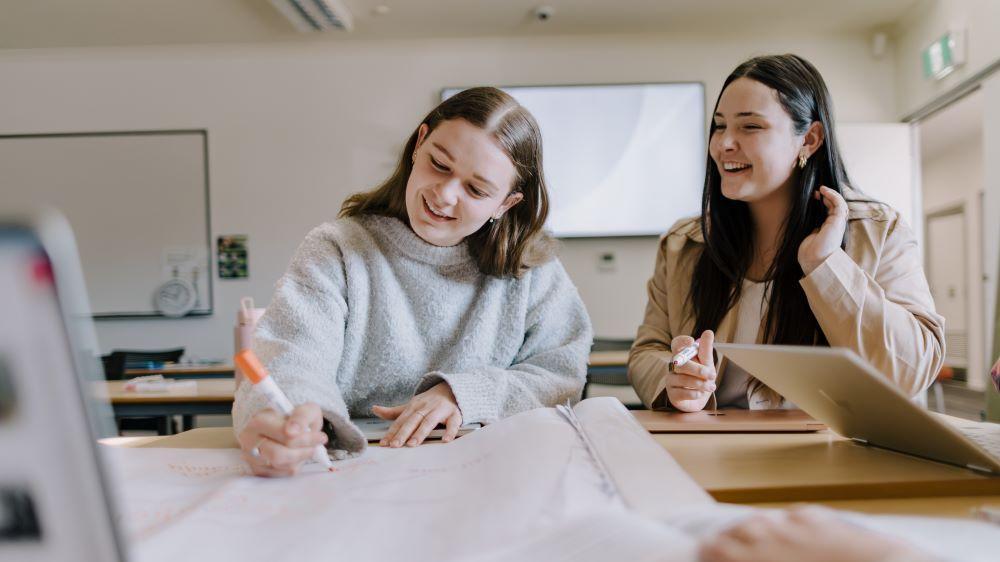 A candid photo of two female UOW students collaborating at a desk. One student, with light brown hair, is focused on writing in a notebook with an orange marker. The other student, with dark hair, smiles and looks on, her hand near her ear. A laptop and a whiteboard are visible in the background.