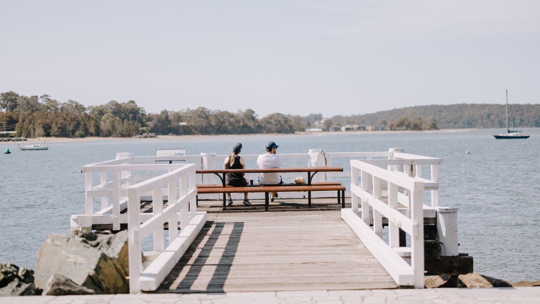 A woman and man rst on a bench at the end of the wharf in Batemans bay