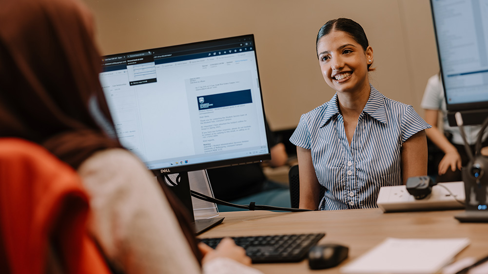 A woman in a striped blouse smiles across a desk in an office setting. A computer monitor with text and a keyboard are in the foreground. The atmosphere is professional and welcoming.