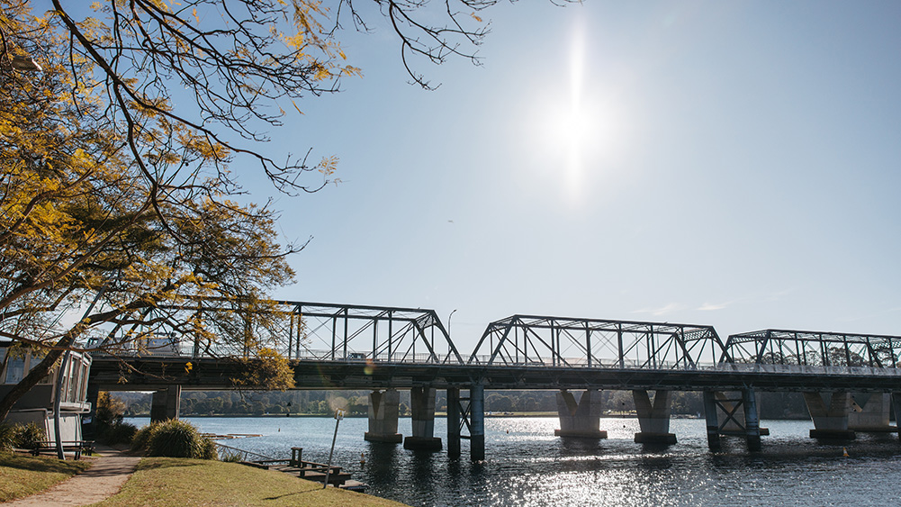 A sunlit iron bridge spans a calm river, framed by autumn foliage and a clear blue sky, evoking a serene landscape.