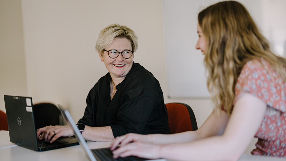 Two women are seated at a table, focused on their laptops, engaged in a collaborative work session in a bright, modern office setting.