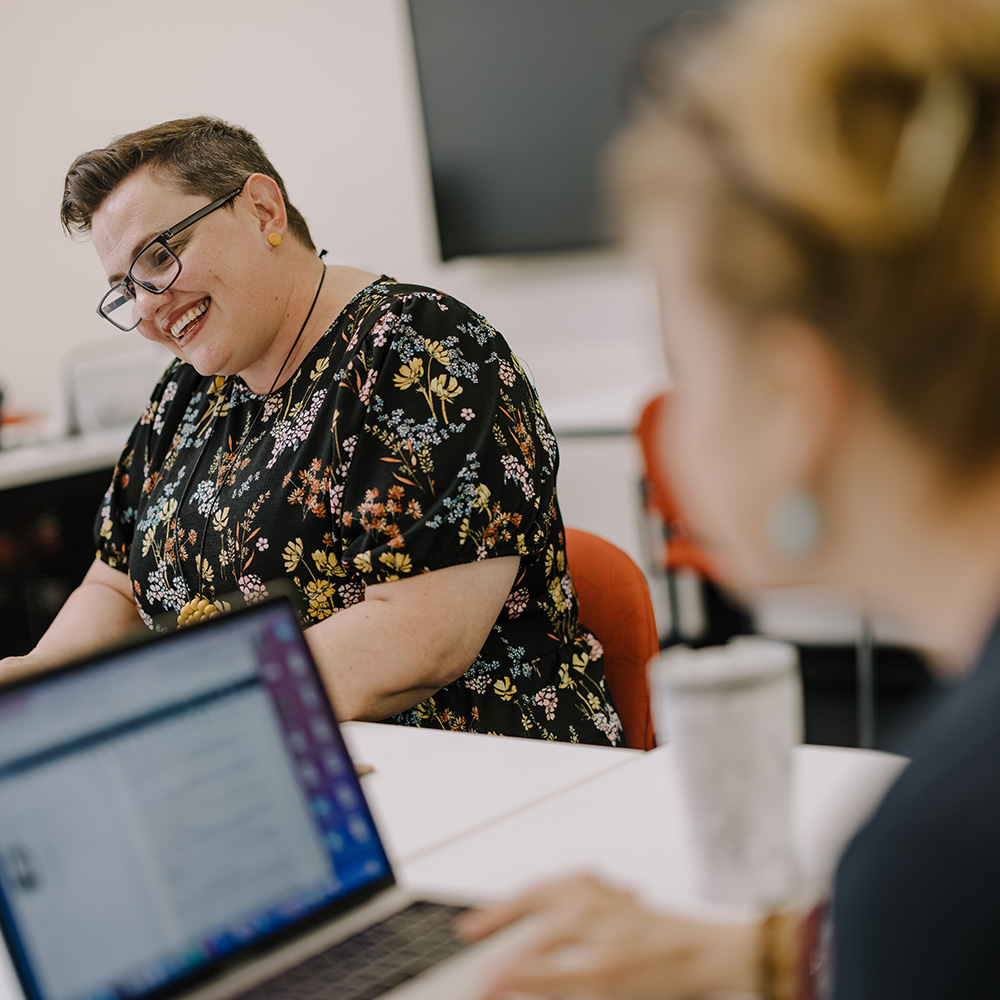 A person in a floral black shirt sits at a table, focusing on a laptop, with another person partially visible in the background.