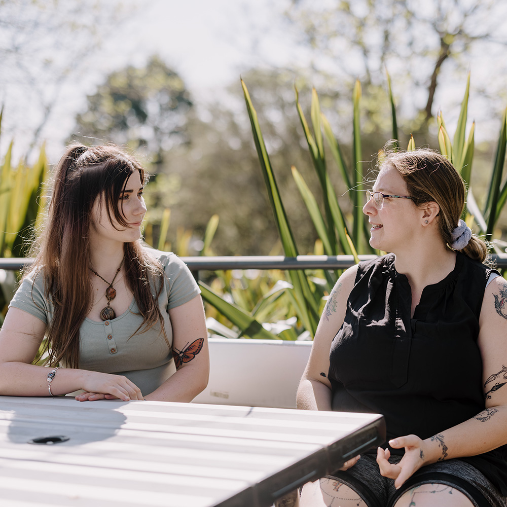 Two women sit at a table outdoors, surrounded by lush greenery. One wears a light green top and the other a black sleeveless shirt.