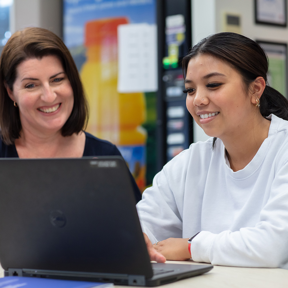 Two women are engaged in a focused discussion over a laptop on a table, set against a colorful background.