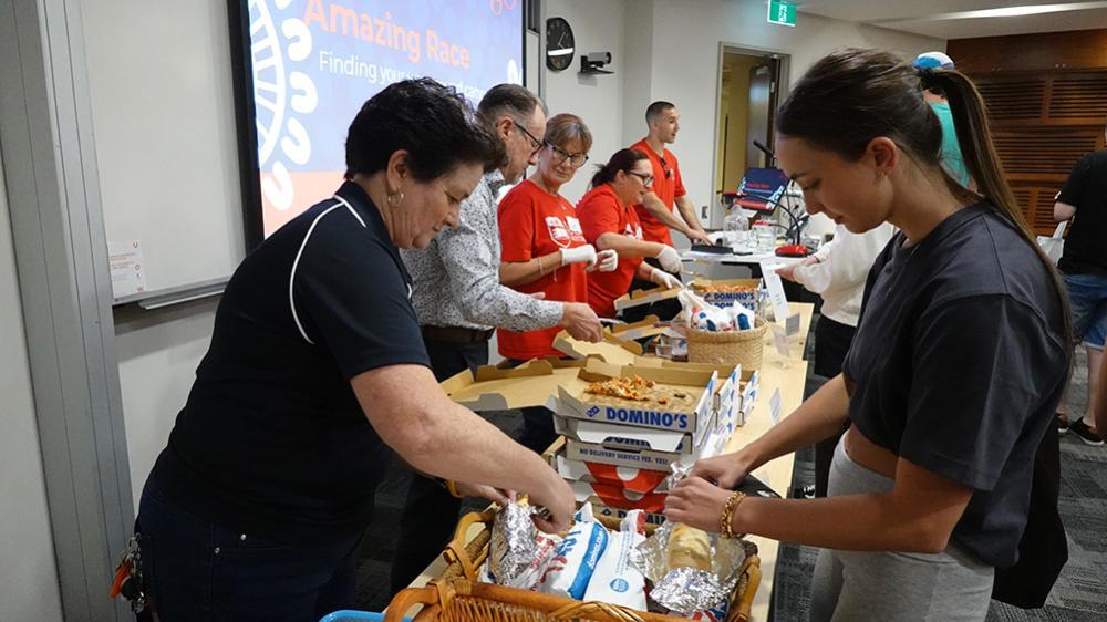 A group of people gather around a table filled with food, including pizza and snacks, in a brightly lit room for an event.