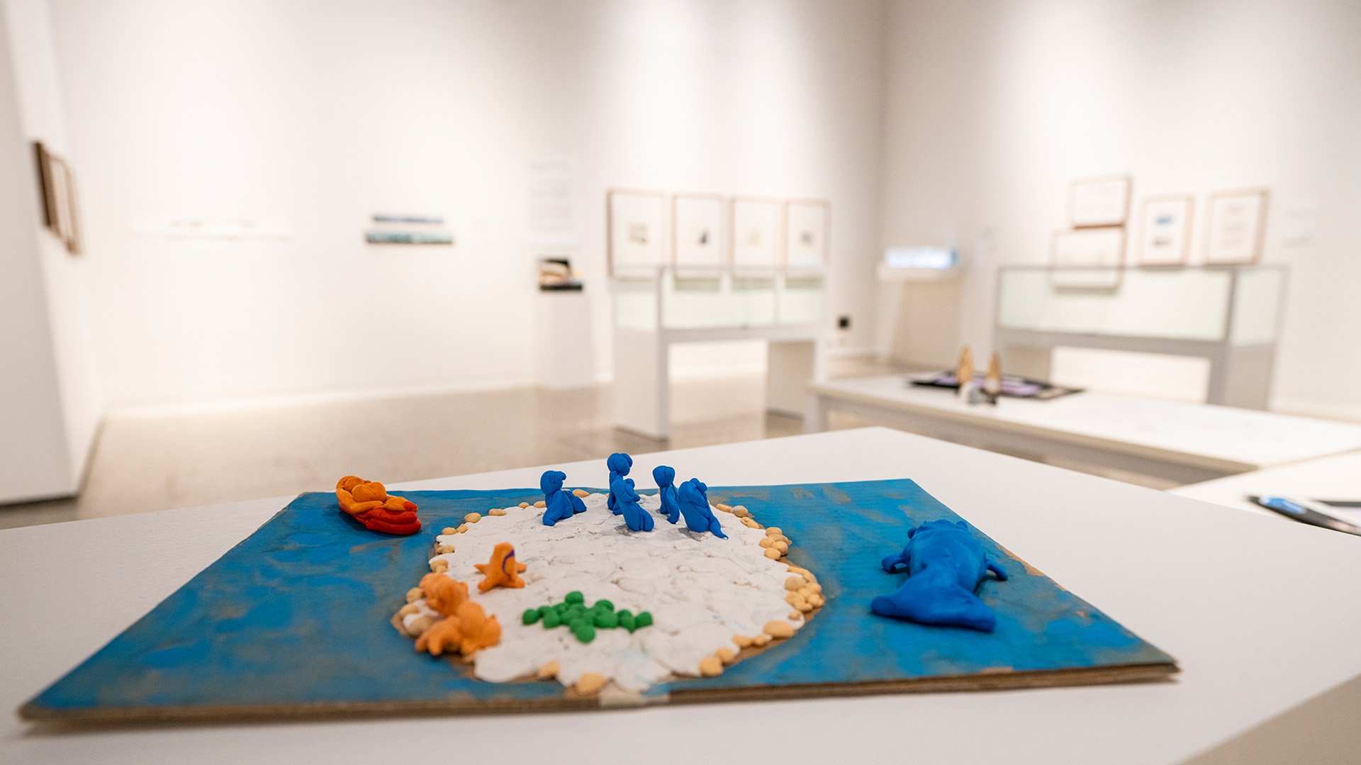 A small clay art piece on a pedestal depicts blue and orange figures on a white island surrounded by blue water. Blurred gallery walls visible behind.