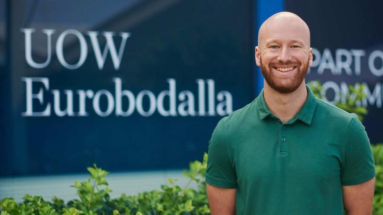 Male wearing green polo shirt, standing in frnot of windows signs that say UOW Eurobodalla smiling at camera