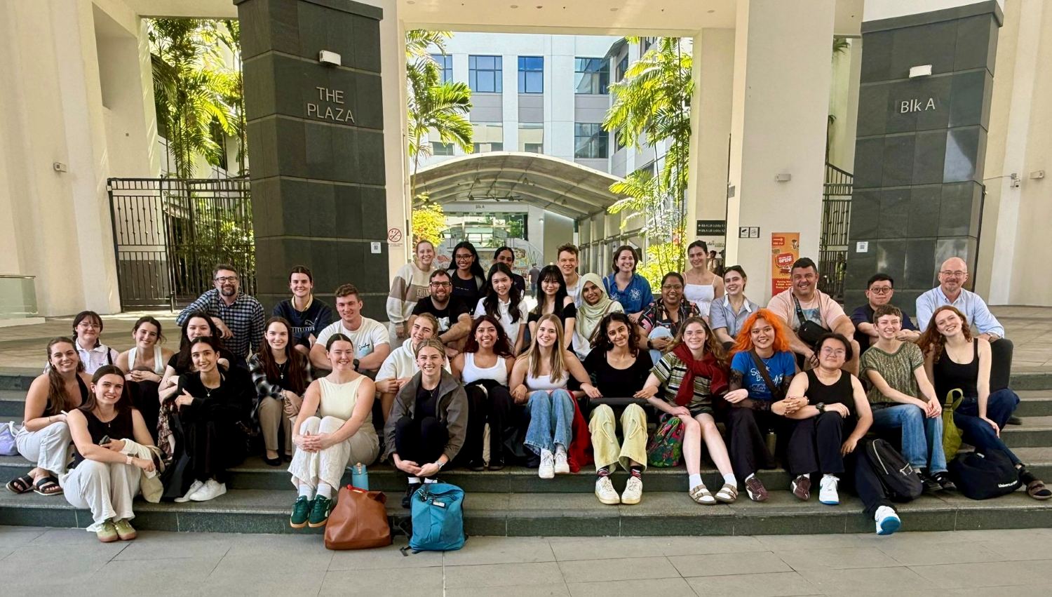 A large group of people sit on the staors at the opening of a hotel called 'The Plaza'