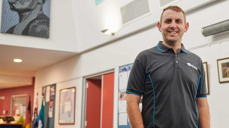 Male in grey shirt, smiling, looking in to camera. Standing in a foyer