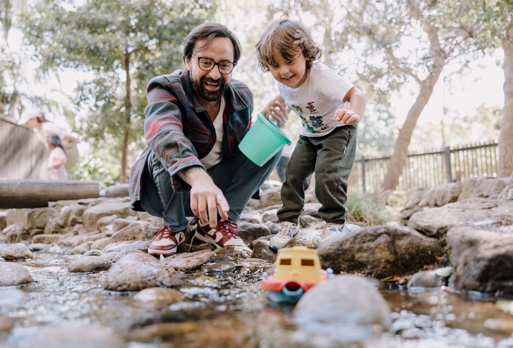 A preschool aged boy is with his Dad, watching a toy boat get pushed downstream by water