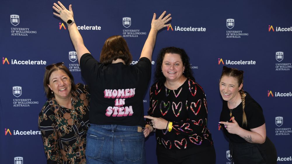 A woman has her hands in the air, her back to the camera, 2 friends on her right,1 on her left. All 3 are smiling and looking at the camera and pointing to her back which says 