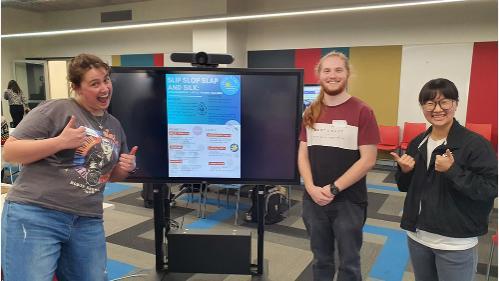 Three people looking happy as they stand beside a presentation screen. One has his thumbs up.