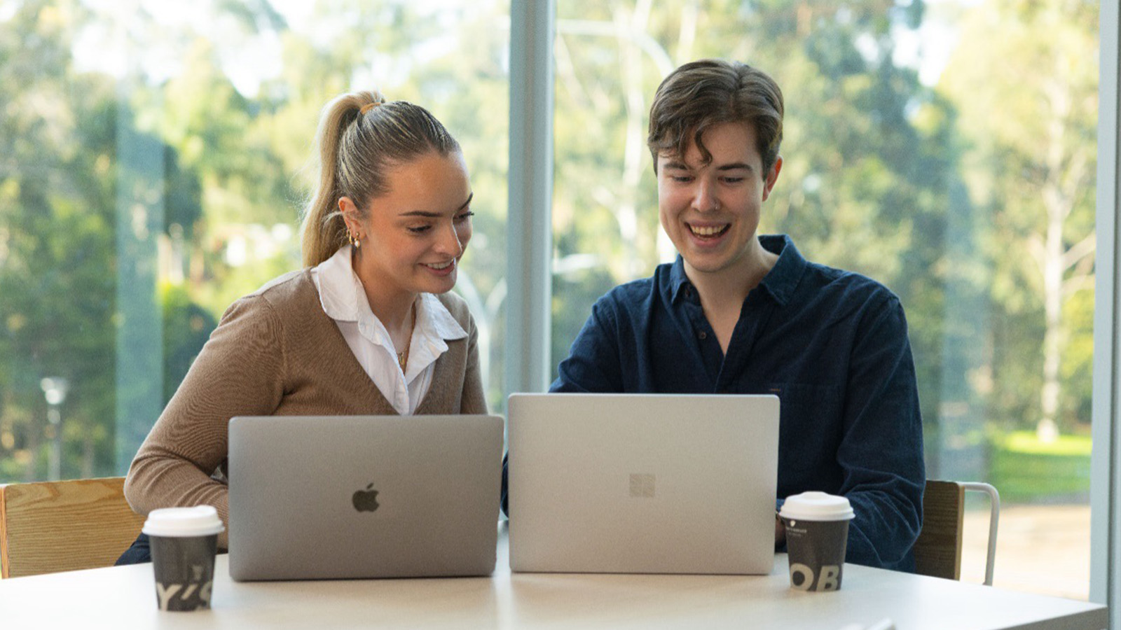 Two student sitting with their laptops at a table collaborating