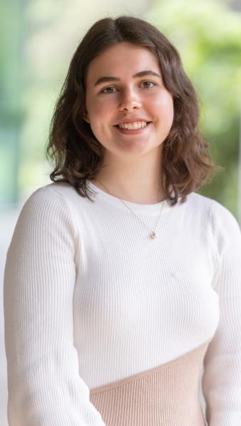 A female student stands smiling at the camera. She is wearing a white knit dress.