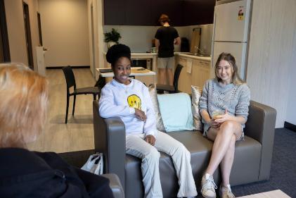 Two students sitting on a lounge talking with student in the background in a kitchen cooking