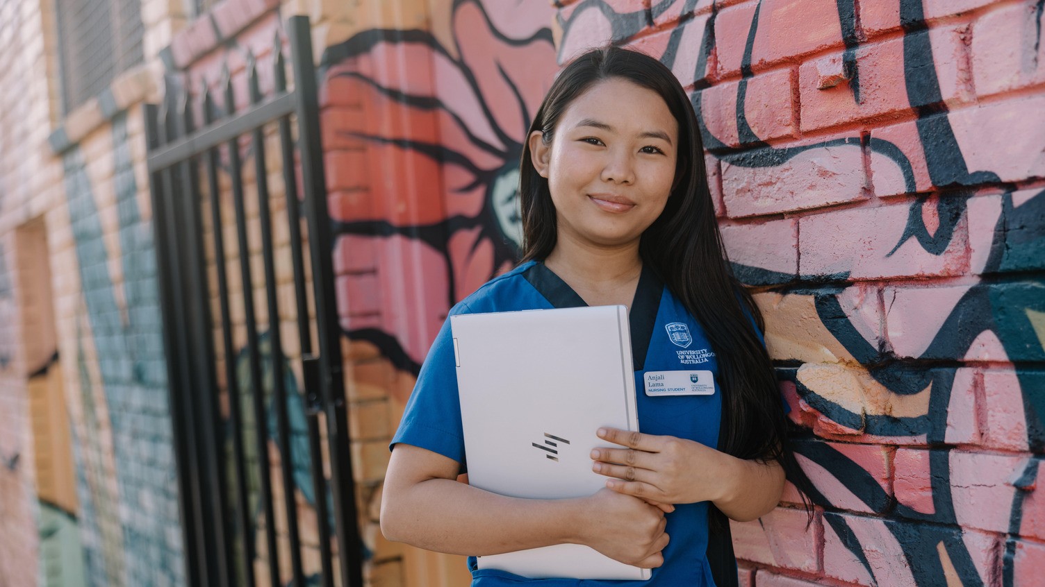Student in nursing uniform holding a laptop beside a colourful mural wall.