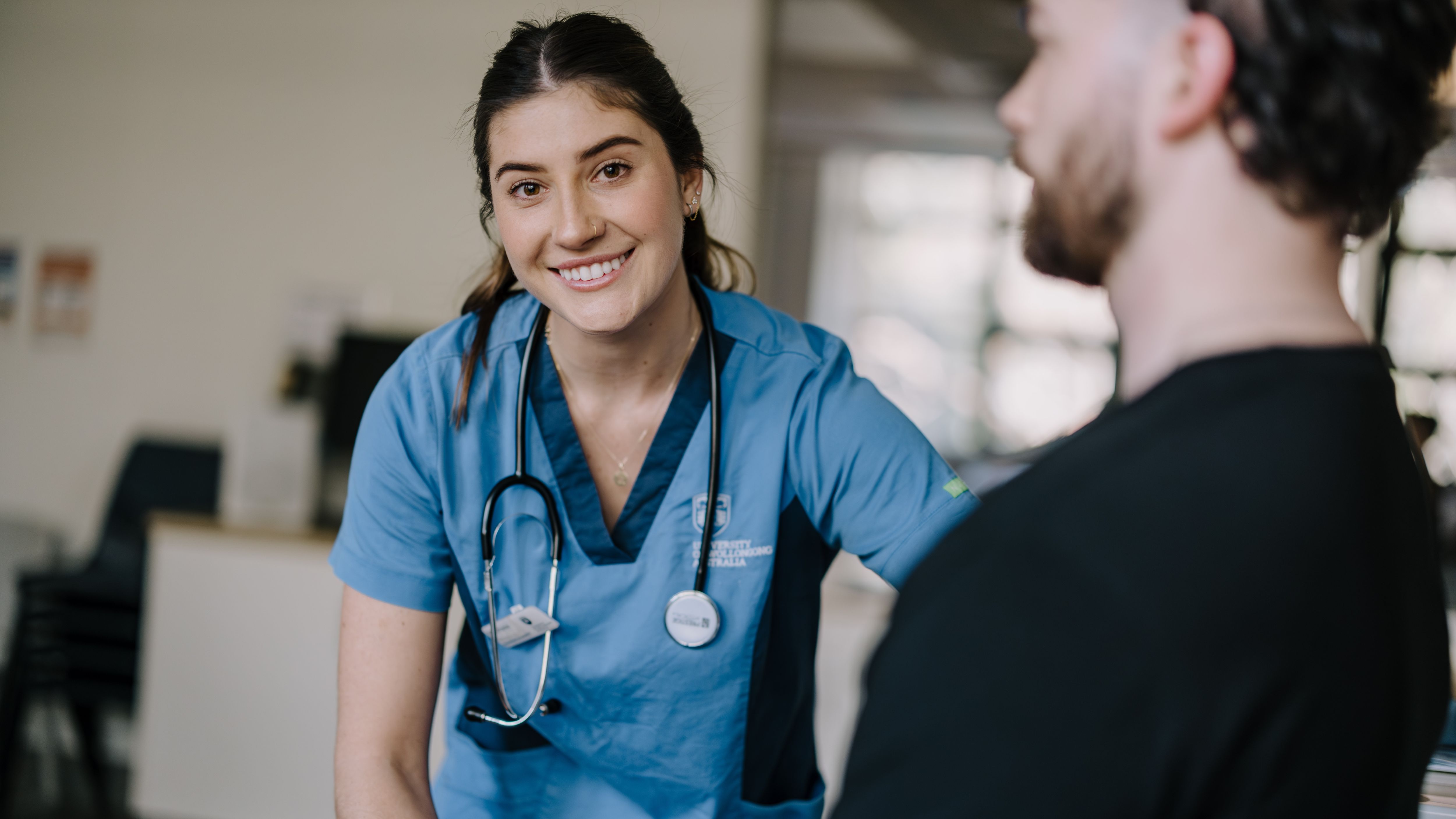 Student nurse in scrubs speaking with a person in a clinical setting.