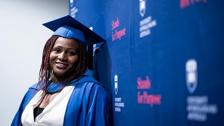 Image of a student dressed in a graduation gown leaning against a wall