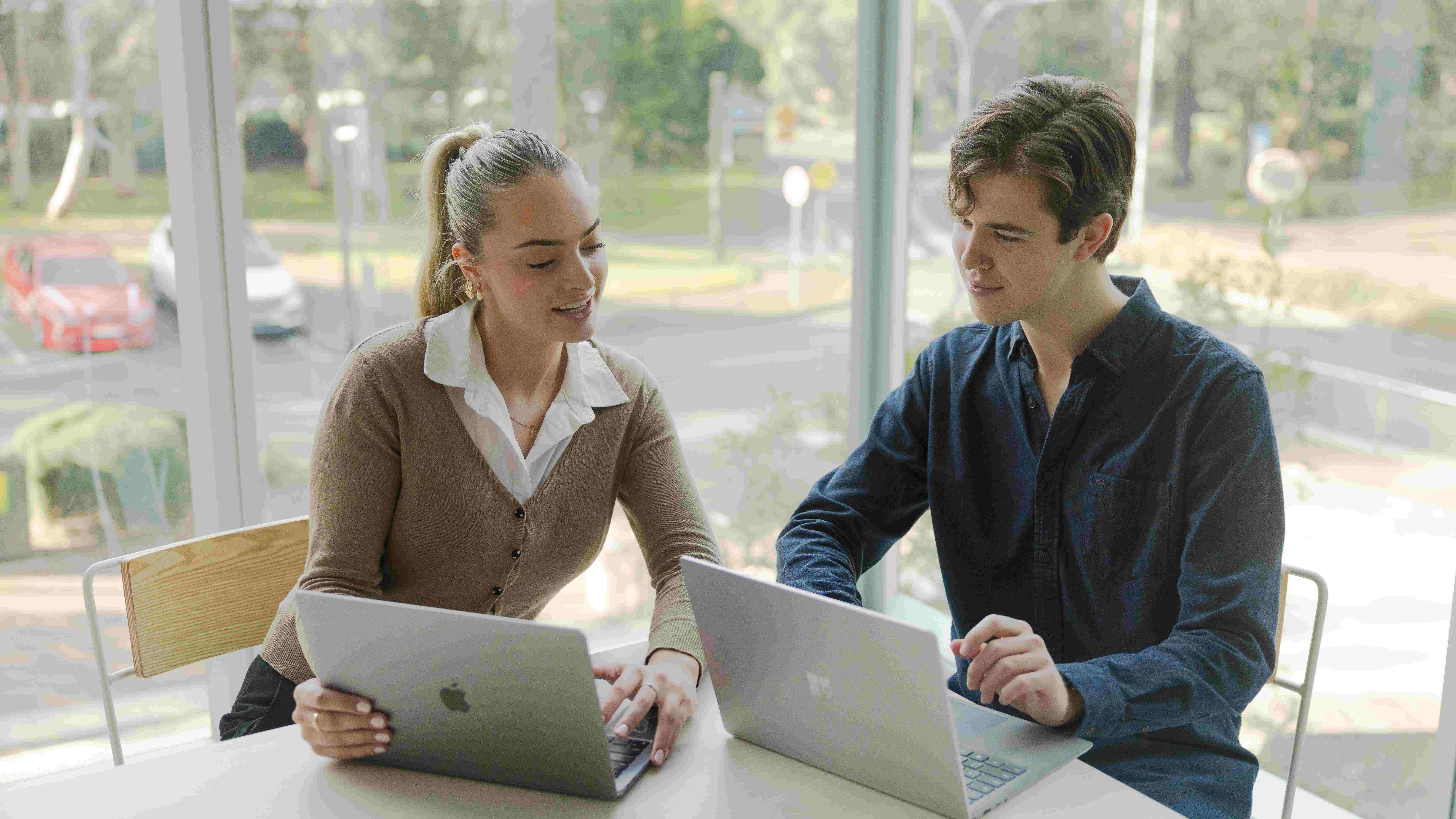 Two people sit at a table by large windows, working on laptops and discussing documents
